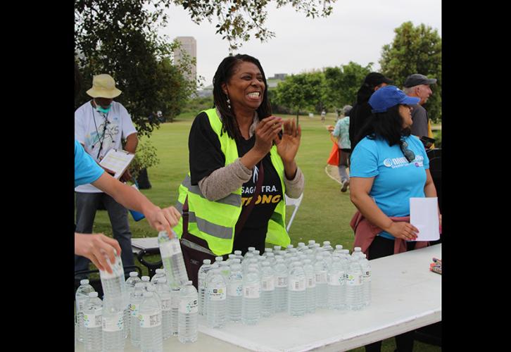 A Black woman in a black “SAG-AFTRA strong” shirt and a safety vest smiles and claps behind a table of water.