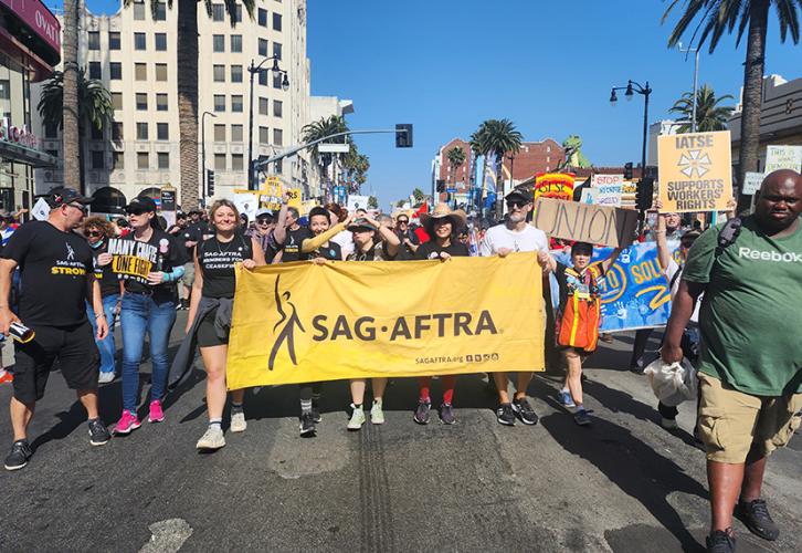 A large group of  people of different ethnicities walk in the street with a yellow SAG-AFTRA banner.