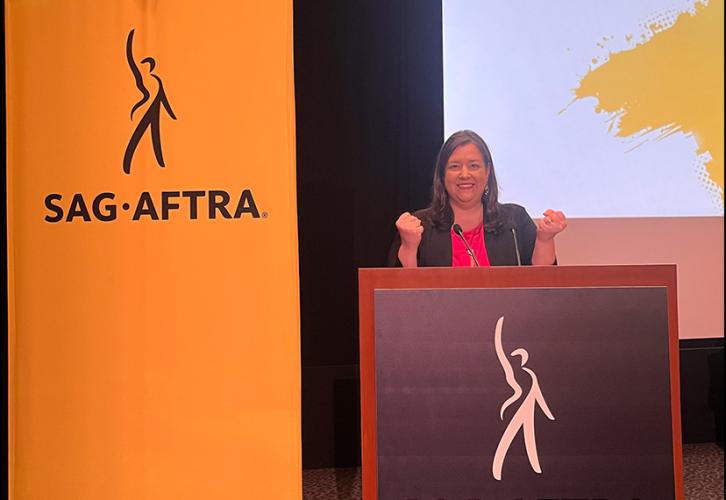 Damon stands behind a podium with the SAG-AFTRA logo. She smiles with her fists clenched. Beside her is a standing yellow SAG-AFTRA banner. 