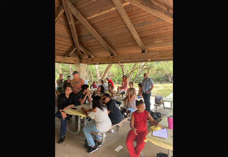 An outside photo of a group of diverse men, women and children staring to the left. Some have plates of food.  