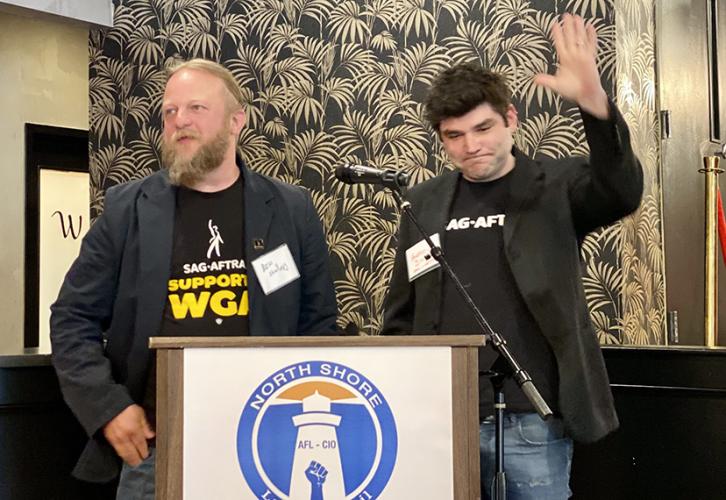 Shallop and Smith, both dressed in black, stand at a podium with an AFL-CIO logo. Shallop looks to his right and Smith gestures with his hand in the air.
