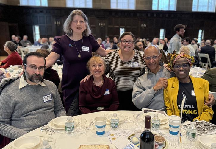 Some sit around a table in a crowded room, while Appel and Stoolmacher stand behind them. James wears a SAG-AFTRA Strong shirt.