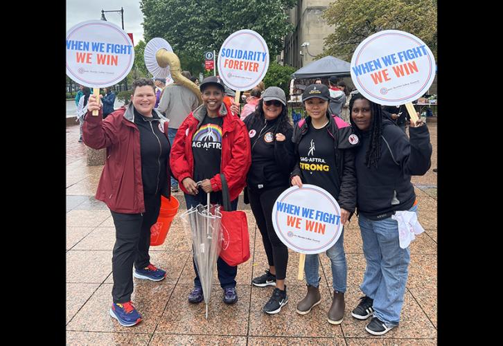 The group stands outdoors, and it appears that it has recently rained. Many hold "When We Fight, We Win" signs. Lyman holds an umbrella.
