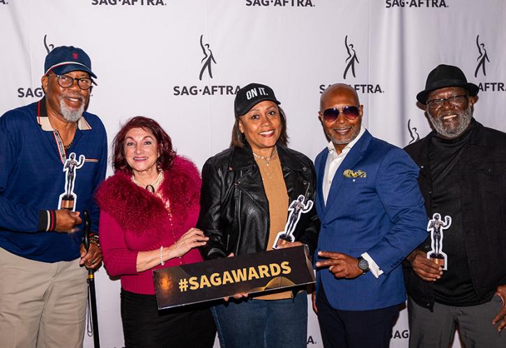 Cinco miembros sonríen mientras posan frente a una pancarta con logotipos de SAG-AFTRA. Tienen réplicas de la estatuilla del Actor y un cartel que dice "#SAGAWARDS".