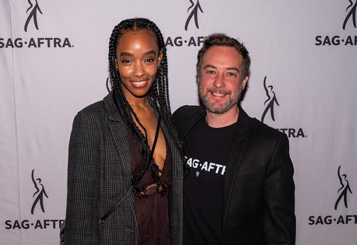 Una mujer y un hombre sonríen mientras posan frente a una pancarta con logotipos de SAG-AFTRA.