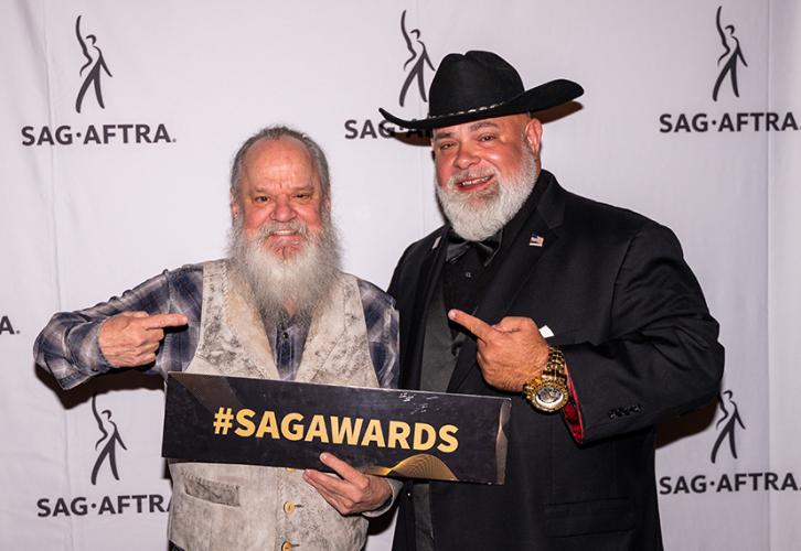Dos hombres sonríen mientras posan frente a una pancarta con logotipos de SAG-AFTRA. El hombre de la izquierda sostiene un cartel que dice "#SAGAWARDS".