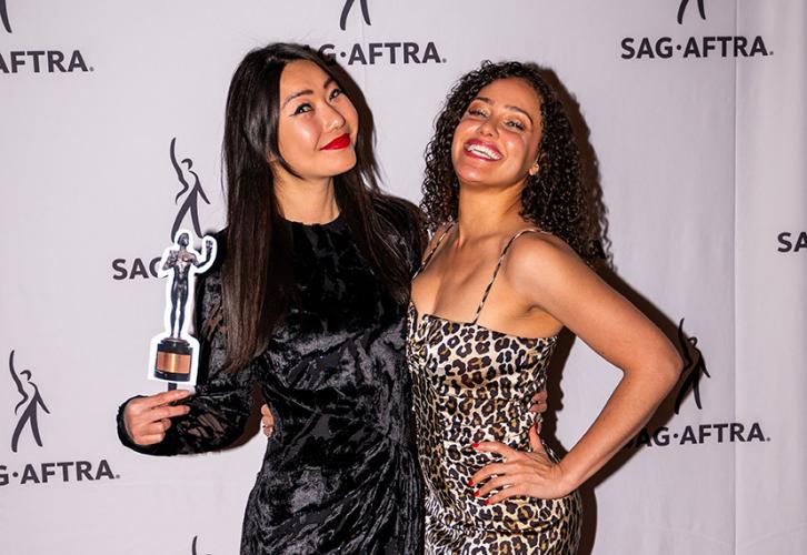Dos mujeres sonríen mientras posan frente a una pancarta con logotipos de SAG-AFTRA. Uno sostiene una réplica de la estatuilla del Actor.
