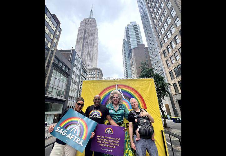 Cuatro personas se paran en una carroza frente a un fondo amarillo con un arco iris. La persona de la izquierda sostiene un cartel con el logotipo de SAG-AFTRA sobre un arcoíris sobre un fondo verde azulado. La segunda persona desde la izquierda sostiene un cartel morado con texto amarillo.