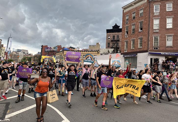 Un gran grupo de personas participa en el contingente SAG-AFTRA de la Marcha del Orgullo de Nueva York. Las personas al frente del contingente sostienen una pancarta amarilla del desfile SAG-AFTRA. Otros tienen carteles de color violeta y verde azulado con el logotipo de SAG-AFTRA y un arcoíris.