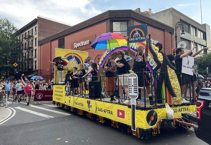 Un grupo de personas monta una carroza amarilla mientras la carroza gira en una esquina de la ciudad de Nueva York. Sostienen paraguas con paneles de arcoíris y carteles de SAG-AFTRA con un arcoíris sobre un fondo verde azulado.