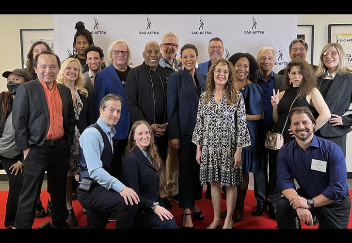 A diverse group of people stand on a red carpet with a white backdrop which reads Sag-Aftra in black text.