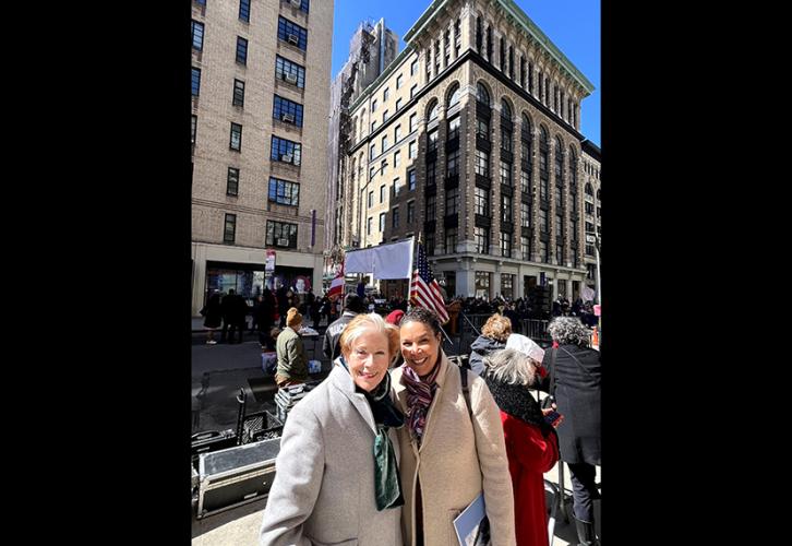 Reardon and Powell smile for the camera. They are standing on the sidewalk in front of a crowd. Buildings and an American flag can be seen in the background.