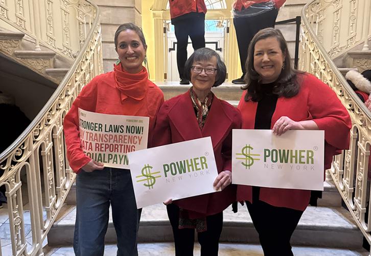 Three people wearing red stand on the steps of City Hall with signs that read “PowHer New York.” 