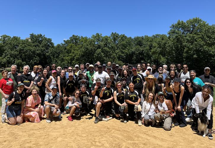 A large group of people gather on a baseball field. There are green trees and a blue sky in the background.