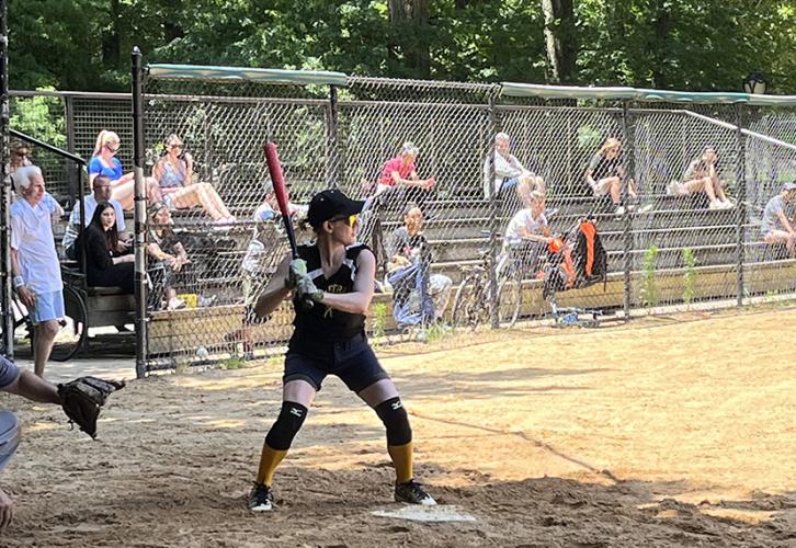 A softball player dressed in black stands at bat with a red baseball bat in her hands.