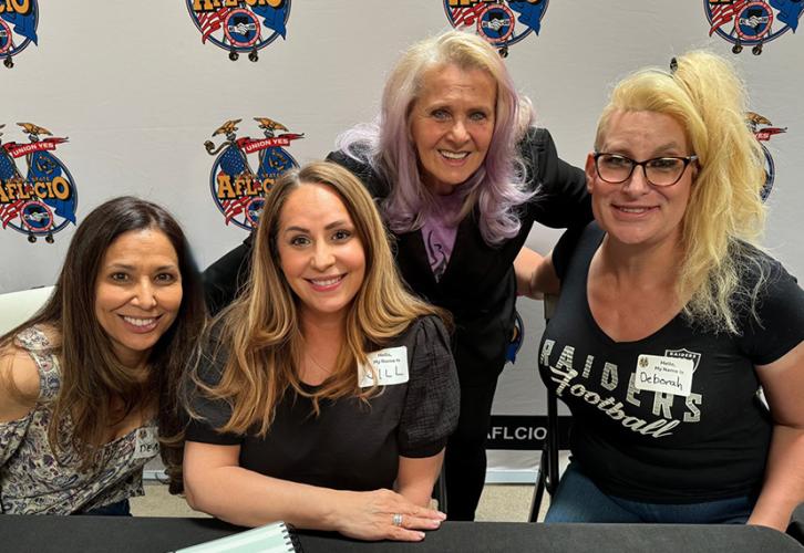 The four women pose in front of a backdrop adorned with AFL-CIO logos. Shapiro wears a multicolored top and the others are in dark clothing.