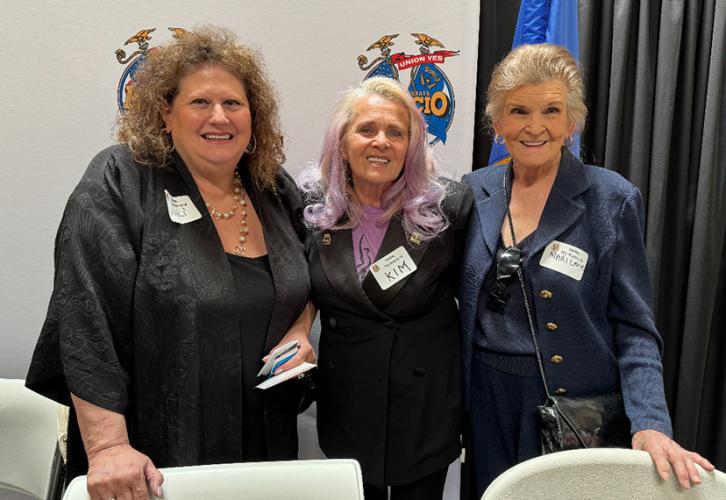 Baskow and Lear rest their hands on chairs in front of them. All wear dark clothing and stand in front of a backdrop adorned with AFL-CIO logos.