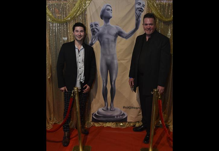 Abellera and Johnathan stand in front of a banner with the picture of The Actor statuette. 