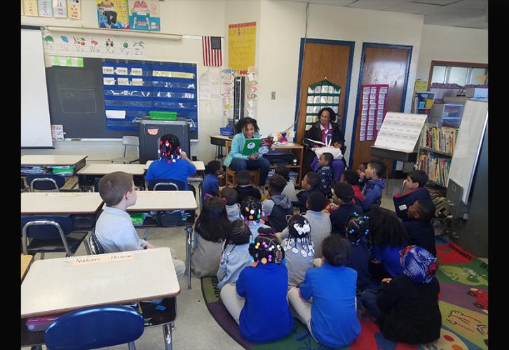 Young African-American girl sits in a rocking chair and reads to children dressed in blue and khaki..