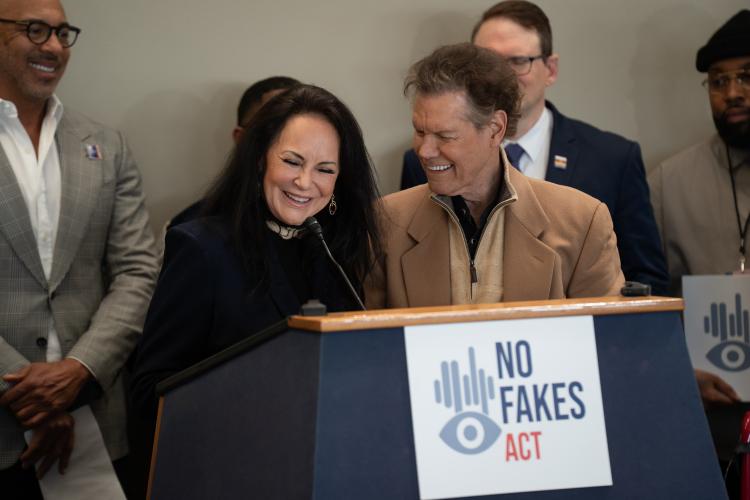 Mary Travis and Randy Travis standing behind a podium adorned with a NO FAKES Act sign.