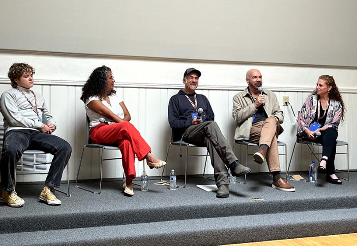Panelists, three men and two women, sit on chairs on a shallow stage. Each holds a mic and Stuart speaks.