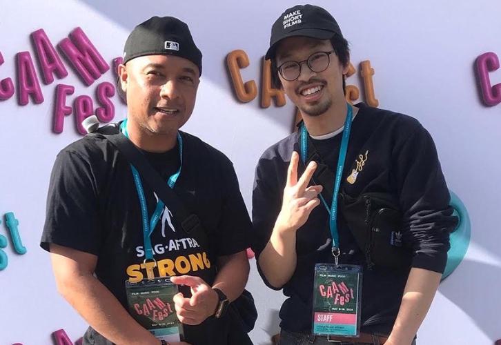 Ison and Hsieh take a photo against a white step-and-repeat backdrop with “CAAMFest” in pink, yellow and gold. Ison wears a black sports cap, a “SAG-AFTRA Strong” T-shirt and jeans; Hsieh wears a black sports cap, black long-sleeved shirt and dark jeans. 