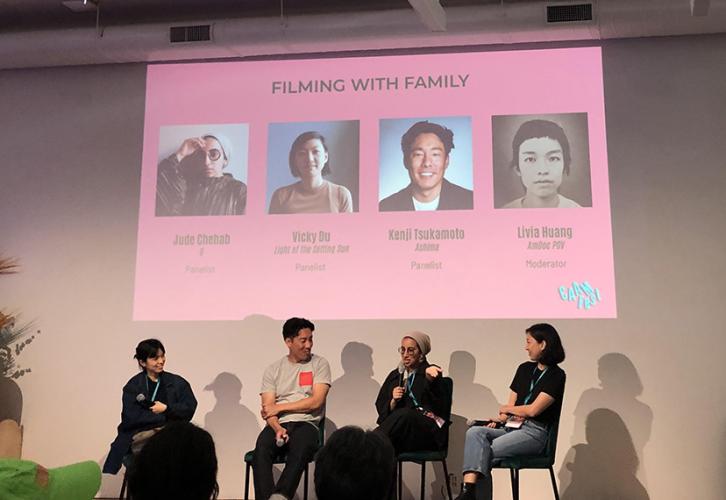 Chehab, Du, Tsukamoto and Huang are all on stage in a room with low lighting. Above them on a projector are their headshots and copy with their names and recent work. Some sound equipment is visible, far right.