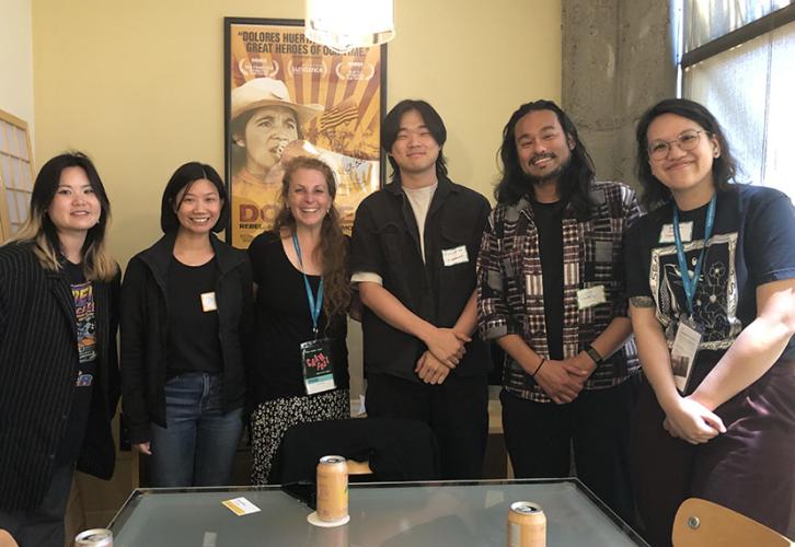 A photo of six individuals, two men and four women, in a conference room setting. Aside from Escobedo, third from left, all are Asian American. They all wear dark clothing. Also visible are a poster on a yellow wall, a window, far right, and a table with 