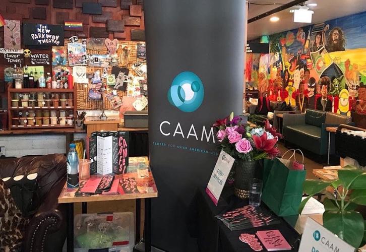 An image of a CAAMFest standing banner, black with an abstract logo of interconnected circles. Framing the banner is festival memorabilia. In the background are walls with colorful artwork and a workshop station with knickknacks.