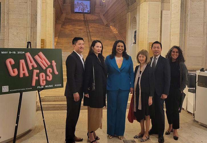 Six individuals stand in a museum lobby, four women and three men. In the center is Mayor London Breed (Black) who wears a distinctive blue pants suit. To the far left is a CAAMFest poster; on the far right is an event staff member (Black, female). 