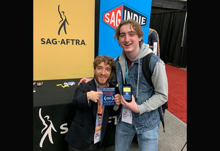 Two men take a photo with pro-union information and a digital membership card in front of the SAG-AFTRA and SAGindie backdrop.  