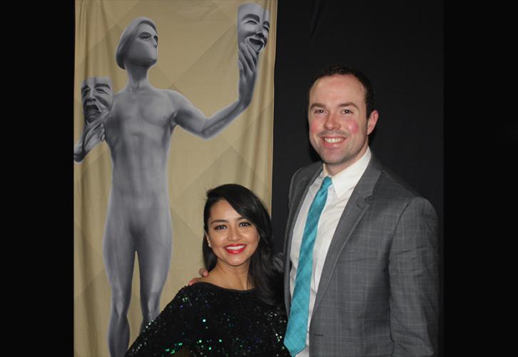 Woman in black sequined dress smiles with taller gentleman in gray suit with award banner in the background.
