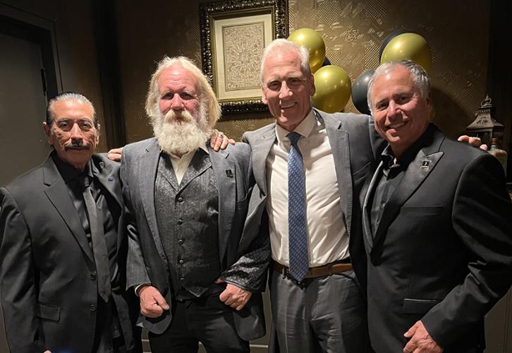 The four gentlemen in suits stand in front of a wall with gold and black balloons. 