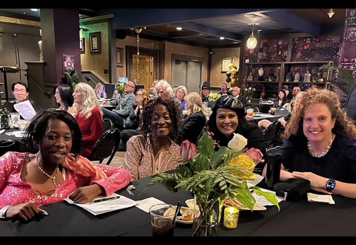 Four women sit at a table. Several other attendees can be seen sitting at tables in the background.