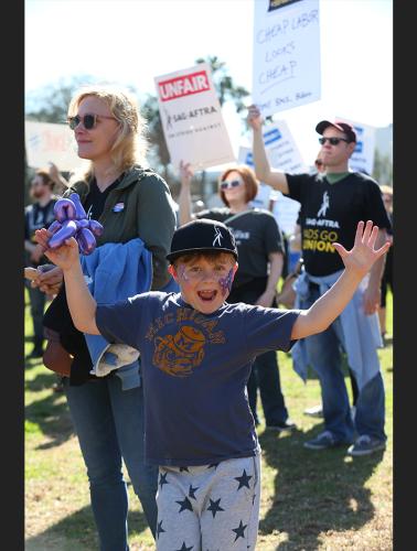 Young boy, holding his hands up enthusiastically wearing a SAG-AFTRA hat and blue t-shirt with grey pants and dark blue stars