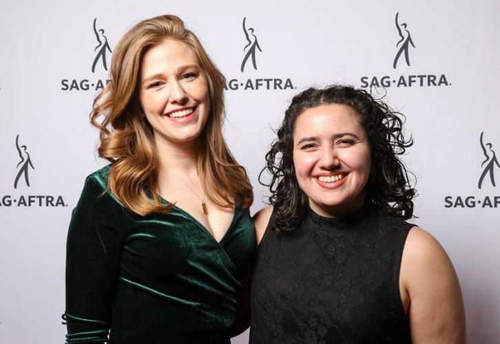 Una mujer rubia vestida de verde se encuentra junto a una mujer de cabello oscuro vestida de negro. Ambos están sonriendo.