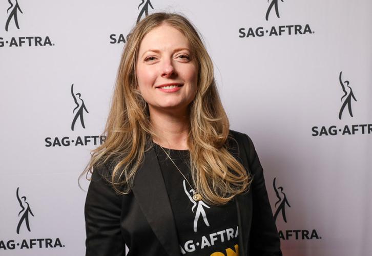 Una mujer con el pelo largo y rubio sonríe. Lleva una camiseta SAG-AFTRA.