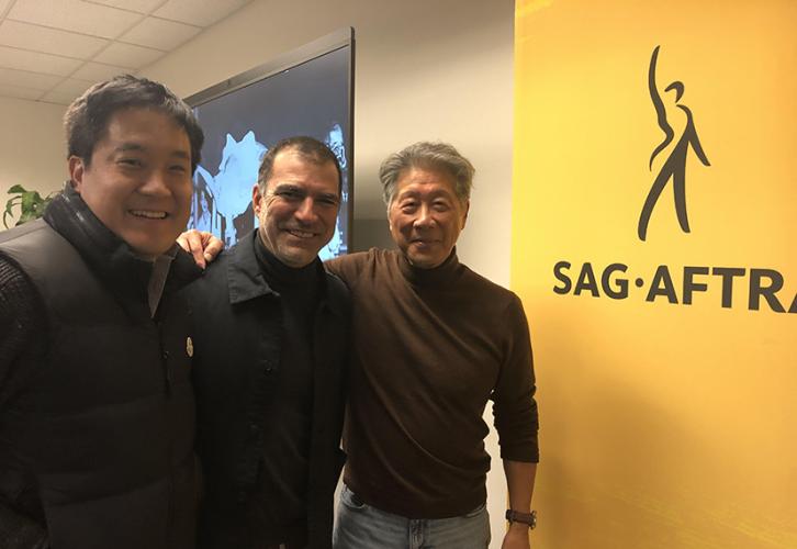 Three people smile and pose for a photograph while standing next to a yellow SAG-AFTRA banner.
