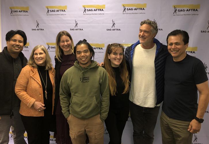 A diverse group of seven people stand together in front of a white step-and-repeat bearing multiple SAG-AFTRA logos.