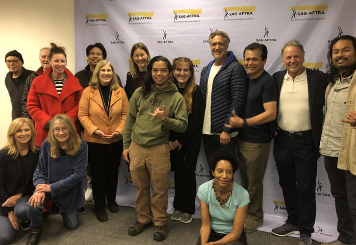 A large, diverse group of people stand together in front of a white step-and-repeat bearing multiple SAG-AFTRA logos. Three others sit on the floor.