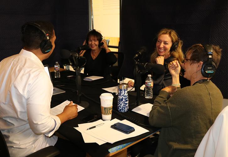 David white sitting at a table with headphones wearing a white button up smiling sitting next to Carteris in a olive Cardigan and headset. Bender and Harcharic sit side by side and across the table from White with headphones on.