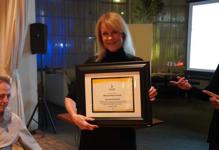 Burkhead holds her framed certificate. On the left there is a seated man; on the right, some unknown persons’ hands appear. 