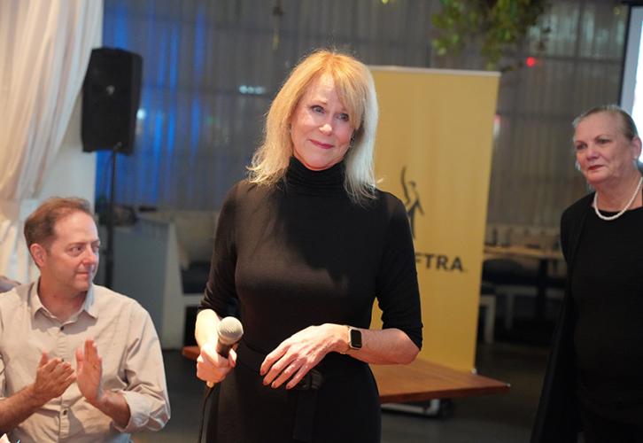 Burkhead stands in the center of the photo. She makes a relaxed gesture with her hands; in her right hand is a microphone. Behind her on the left is a man who is clapping and looking towards his left; on the right, a woman looks on. Visible in the background is a standing yellow banner with SAG-AFTRA’s logo.