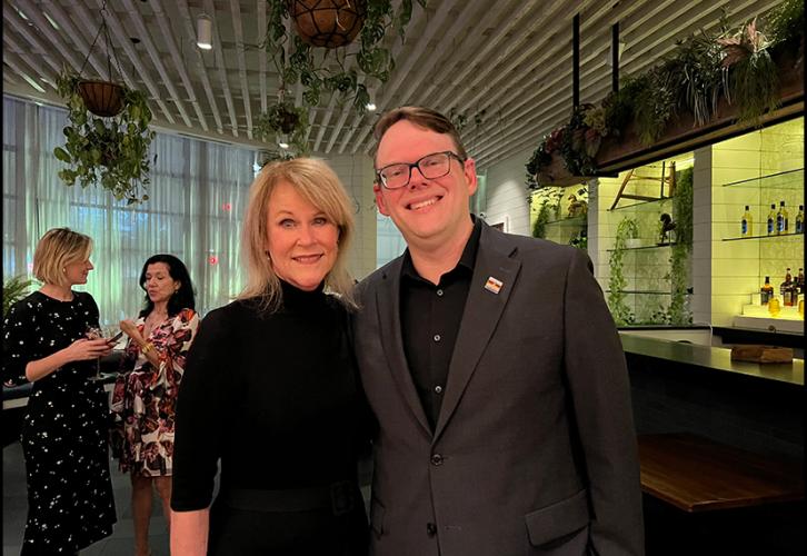 Burkhead and Crabtree-Ireland stand closely together in what is a bar or restaurant-like setting. Crabtree-Ireland wears a dark blazer with a SAG-AFTRA enamel pin and black dress shirt. Behind them, two women have a conversation. Also visible are bar and restaurant decor and plants.