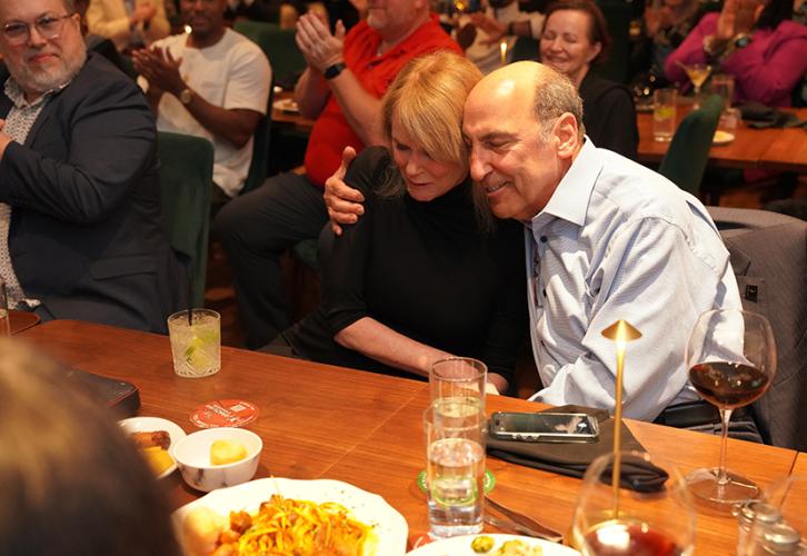 A woman and a man, seated at a restaurant table, embrace. Around them, others applaud. On the table are plates of food and glasses. 