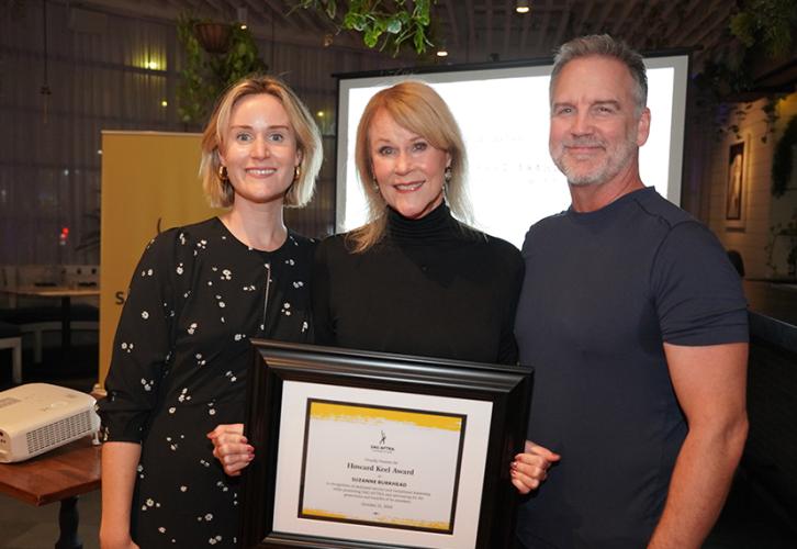 Maggie Burkhead, Suzanne Burkhead and Anderson stand closely together. Maggie Burkhead wears a black dress with patterns of small daisies; Anderson wears a dark charcoal shirt. Burkhead holds her framed certificate. Behind them is a blank projector; over Maggie’s shoulder is a yellow standing SAG-AFTRA banner. 