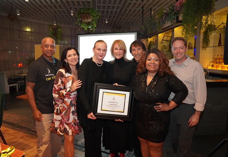 Seven individuals stand closely together in a bar or restaurant-like area. Burkhead, center, holds her framed certificate. Behind the group is a flatscreen with a blank white screen. Some restaurant decor and plants are also visible. 