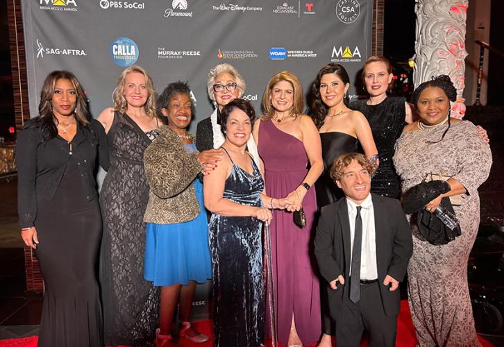 Ten individuals, one man and nine women, stand on a red carpet. All wear formal wear, the man wearing a black suit and tie and white dress shirt, and the women wearing dresses. Behind them is a black step-and-repeat backdrop with logos for SAG-AFTRA, the Media Access Awards, PBS SoCal and others.