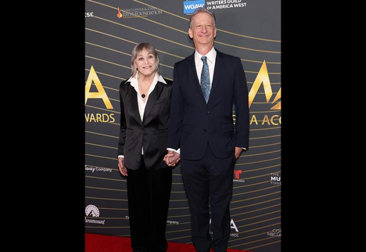 Jewell and Zimmerman stand on the red carpet. They are holding hands. Jewell wears a black suit with a white dress shirt that is slightly unbuttoned, and a necklace with a dark pendant. Zimmerman wears a dark blue suit, white dress shirt and blue necktie. Behind them is a step-and-repeat backdrop for the Media Access Awards.