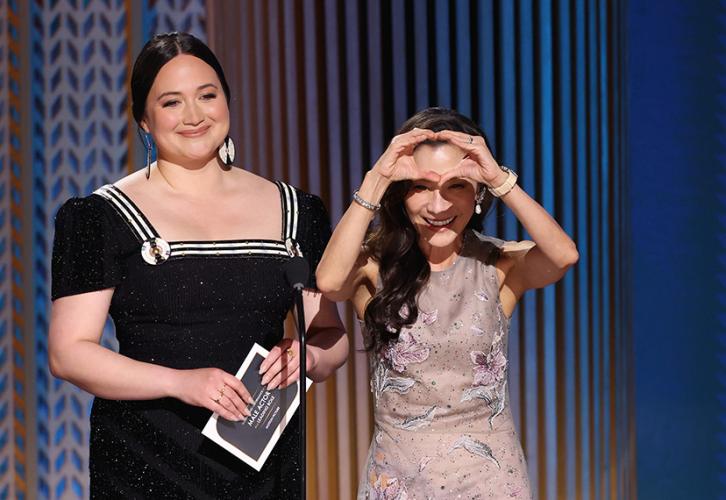 Lily Gladstone and Michelle Yeoh stand onstage. Yeoh holds up her hands in a heart shape before her face.